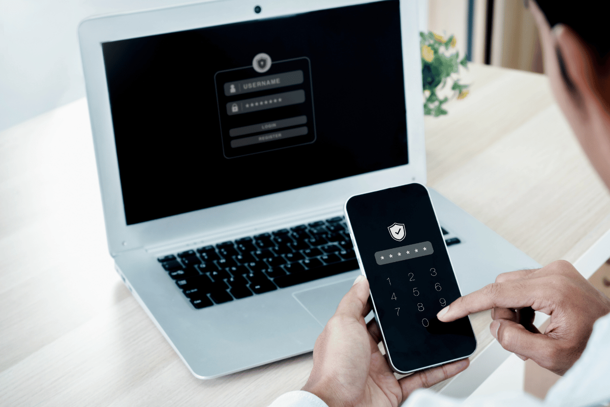 Man on a laptop entering a passcode on his smart phone. 